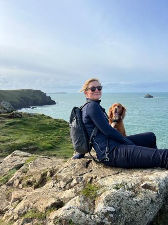 a woman sitting on a rock with a dog, is turning and smiling to the camera. with the sea in the background