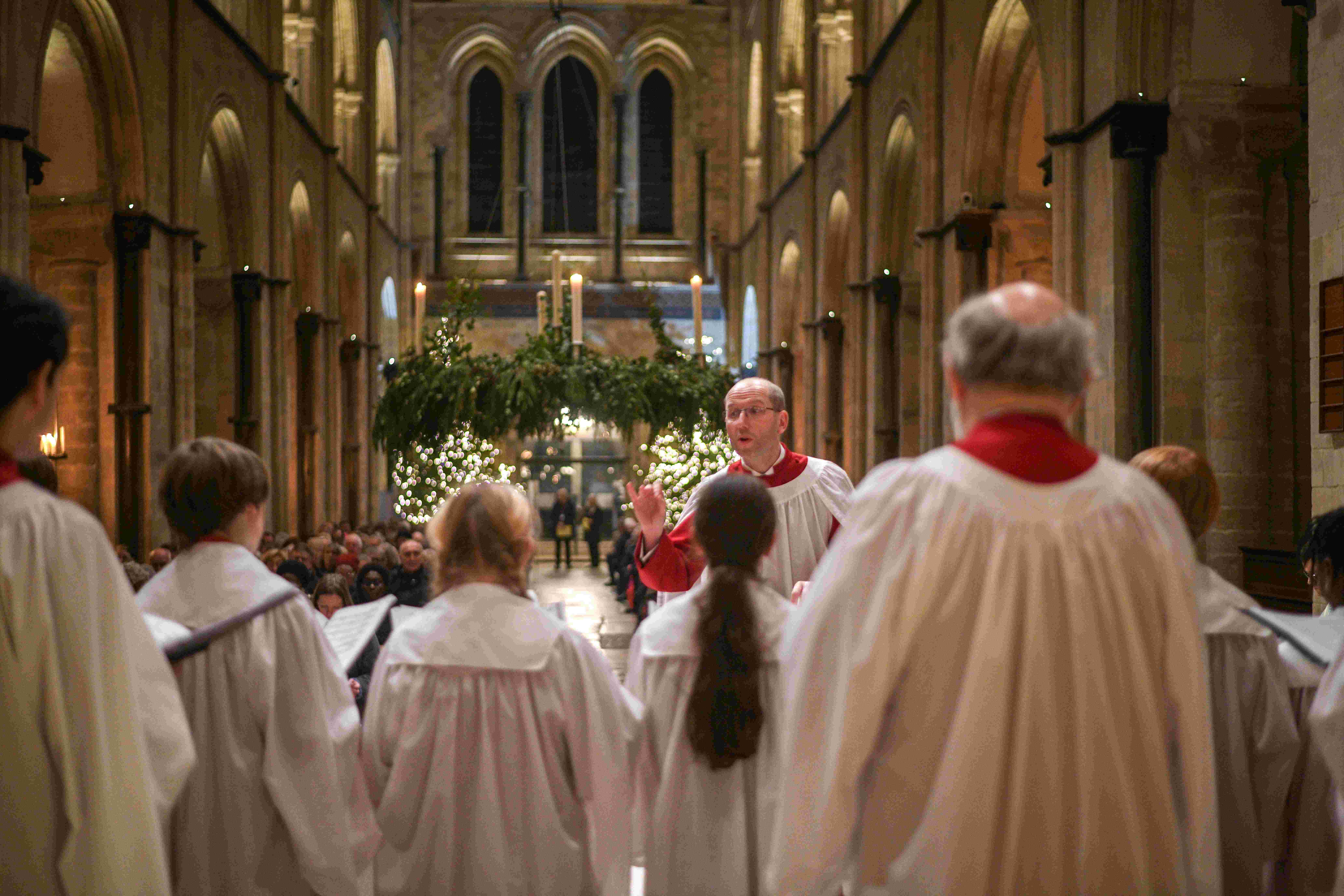 In the foreground are adult and child choristers in white robs, mid ground is a man in white choral robes conducting, in the background are people sitting in seats and above them Christmas foliage