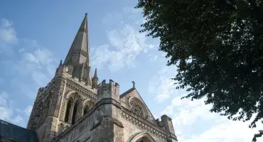 Chichester Cathedral's spire, grey stone, dominates a blue sky. A leafy green tree borders the image which is shot from the ground nearby.