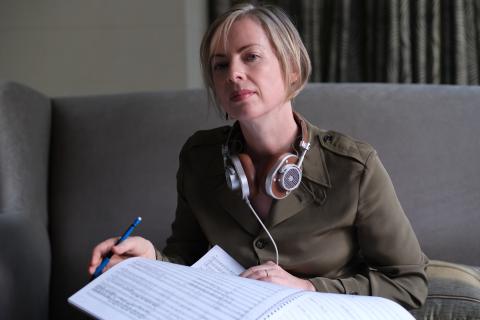 Joanna Marsh, pictured with headphones around her neck and holding sheets of music. Marsh sits on a grey sofa, wearing a green jumper. She has short, blonde hair.