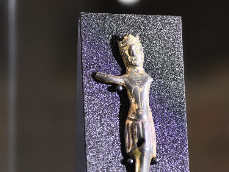 A worn, gold figure setting against a navy blue panel. The figure, a depiction of Christ, wear a crown. Both arms have been damaged - the left completely missing, and the right partially remaining. The figure is no taller than 15cm in height.