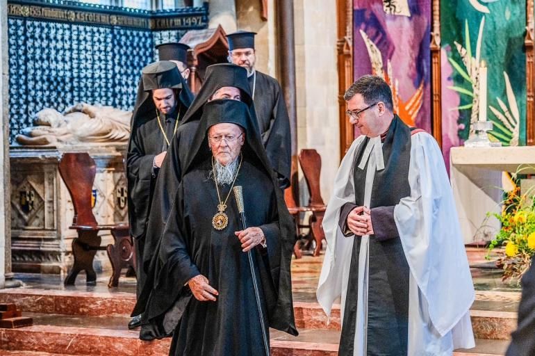 His All Holiness Ecumenical Patriarch Bartholomew walks through Chichester Cathedral accompanied by clergy, greeted by the Dean of Chichester. The Patriarch wears traditional black robes. The group walk in front of John Piper's multicoloured tapestry.