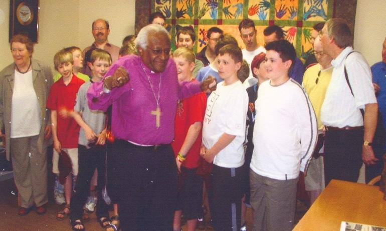 An older black man in purple shirt and wearing a crucifix is dancing in front of a group of children and adults who are looking at him