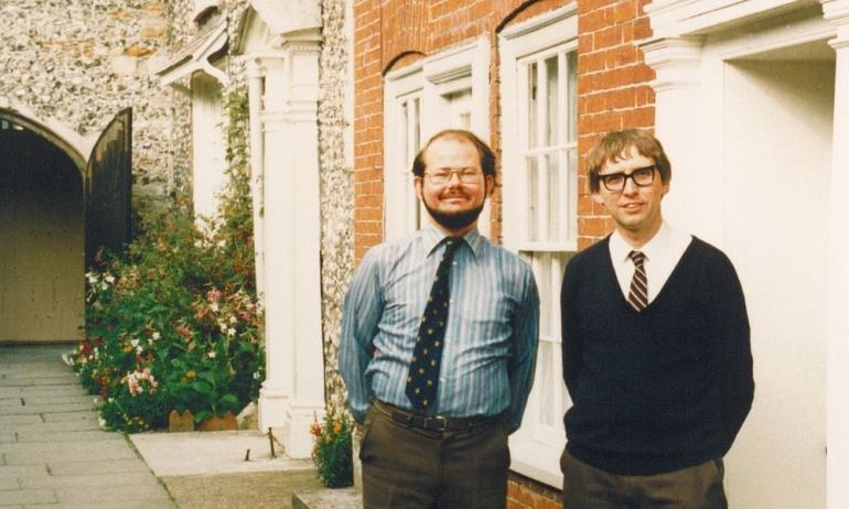 Two white men in late 80s clothing standing in front of a red brick building
