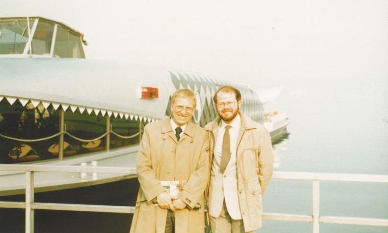 Two white men in 80s clothing standing in front of a boat and water looking to camera 