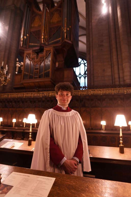 A young man in choir robes stands in front of the pipes of a cathedral organ