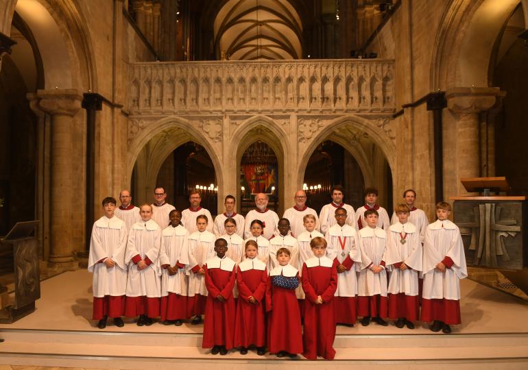 A Choir in red and white robes standing in front of cathedral architecture facing the camera.