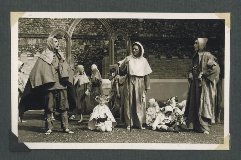 B&W photo of a medieval pageant with people dressed in servant/labourer costumes with the Bishop's Palace in the background
