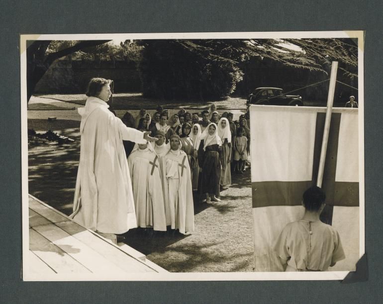 B&W photo of a medieval pageant with people dressed in clerical or servant costumes looking at a robed man on a platform