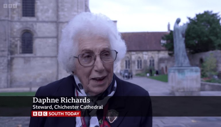 Screenshot with BBC South Today branding of a woman with white hair and wearing black in front of statue and Cathedral