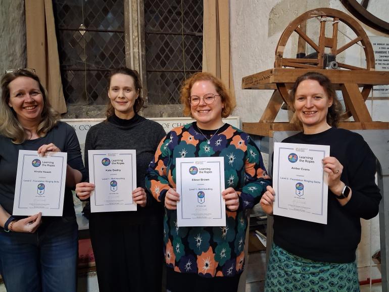 Four individuals standing indoors in front of a stone wall and a tall, arched window inside a historic building. Each person is holding a printed certificate at chest height. To the right, part of a wooden bell‑ringing training wheel is visible. The group is described as bell ringers from Chichester Cathedral.