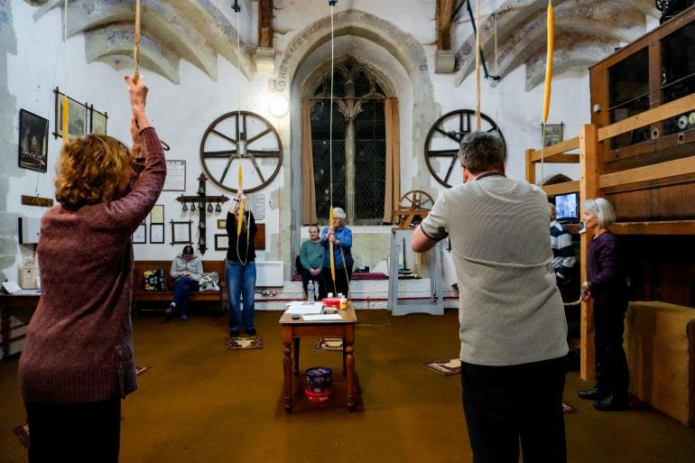 A group of people arranged in a circle inside the bell‑ringing chamber each hold a bell rope raised toward the ceiling. 