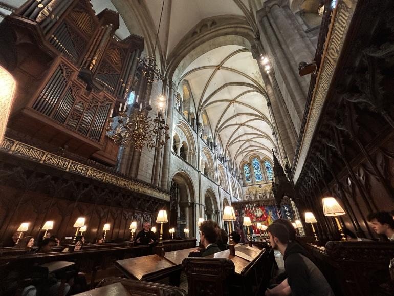 An angled view inside the choir stalls of Chichester Cathedral shows people seated in dark carved wooden seating lit by small individual lamps. The tall vaulted ceiling, ornate pipe organ and stone arches rise above, with a colourful tapestry visible in the distance at the far end of the cathedral.
