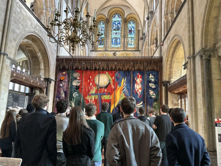 Inside Chichester Cathedral, a group of people stand in front of a large, brightly coloured tapestry behind the altar. The tapestry includes abstract shapes in red, blue and yellow. Above it, tall arched windows and an ornate chandelier hang beneath the vaulted stone ceiling.