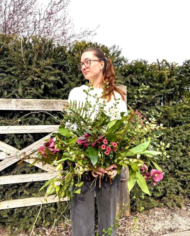 Outdoors near a wooden gate and tall hedges, Nancy Hersey, a former Plumpton College student holds a large arrangement of mixed foliage and flowers. The bouquet includes abundant greenery, long branches, and deep pink blooms, creating a loose, natural display.