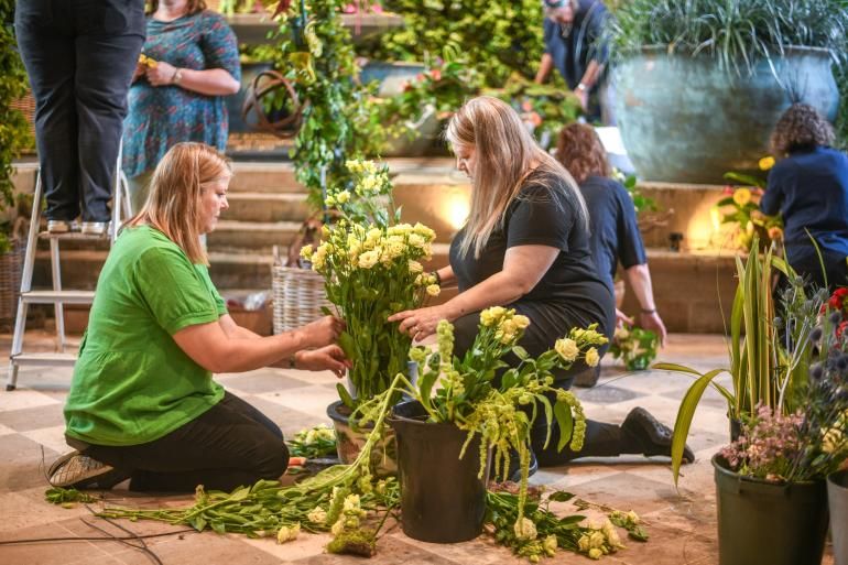 Two people kneel on the floor inside Chichester Cathedral, arranging pale yellow flowers in a large container, with buckets of greenery and cut stems spread around them. In the background, several others work on floral displays among stone steps, large planters, and abundant foliage during the Festival of Flowers 2024